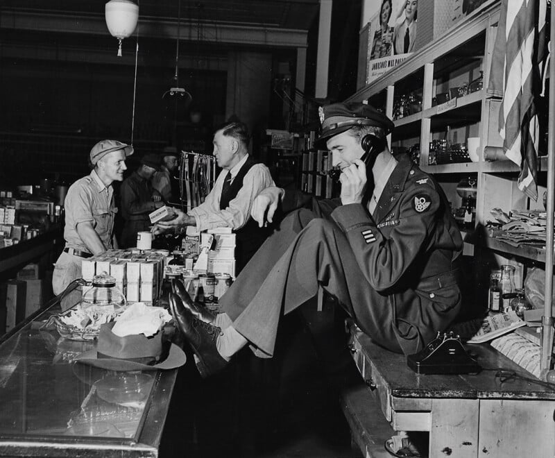A man in a military uniform sits on a counter, talking on a phone, while two men behind the counter interact. Shelves with goods and posters are visible in the background.