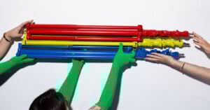 Four hands, two in green gloves, hold three folded metal stands in red, yellow, and blue against a white background.