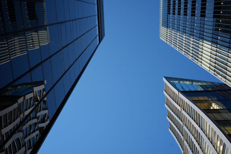 Three modern glass and steel skyscrapers are photographed from below, converging towards a clear blue sky, creating a geometric and symmetrical composition.
