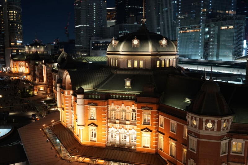 Night view of a historic red-brick building with ornate architecture, illuminated by warm lights, surrounded by modern glass skyscrapers and city lights in the background.