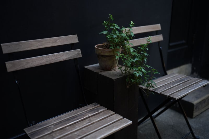 Two wooden chairs sit next to a small, rustic potted plant placed on a wooden block, set against a dark wall in a minimal, moody outdoor setting.