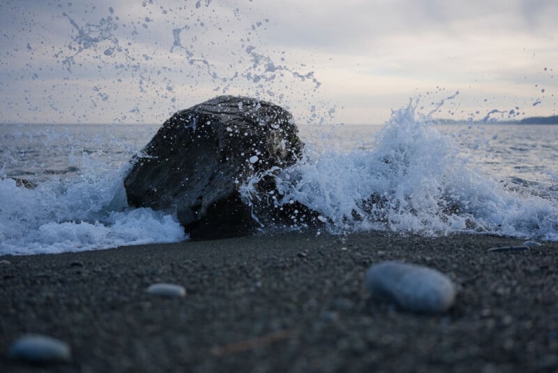 Waves crash dramatically against a large rock on a pebbled beach, sending water droplets into the air under a cloudy sky at the edge of the sea.