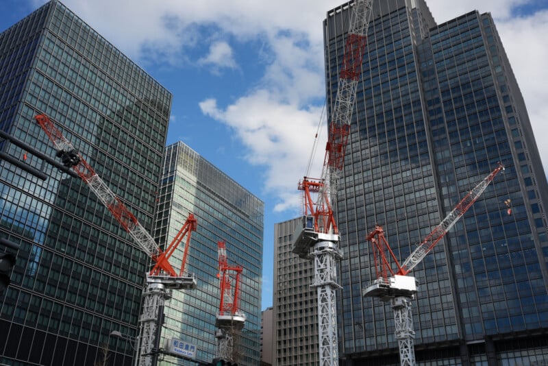 Red and white construction cranes stand tall among modern glass office buildings under a partly cloudy blue sky, indicating an urban area undergoing development.