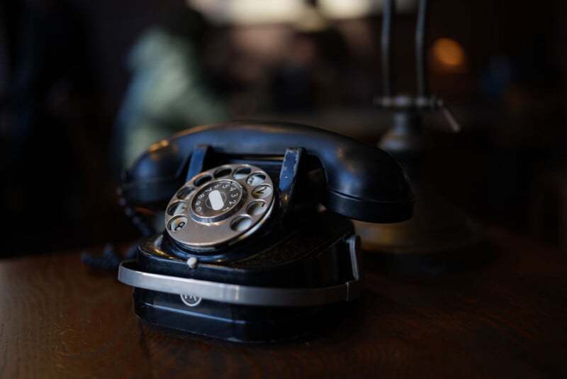 A vintage black rotary telephone sits on a wooden table in a dimly lit room, with a blurred background and soft lighting highlighting the details of the phone.