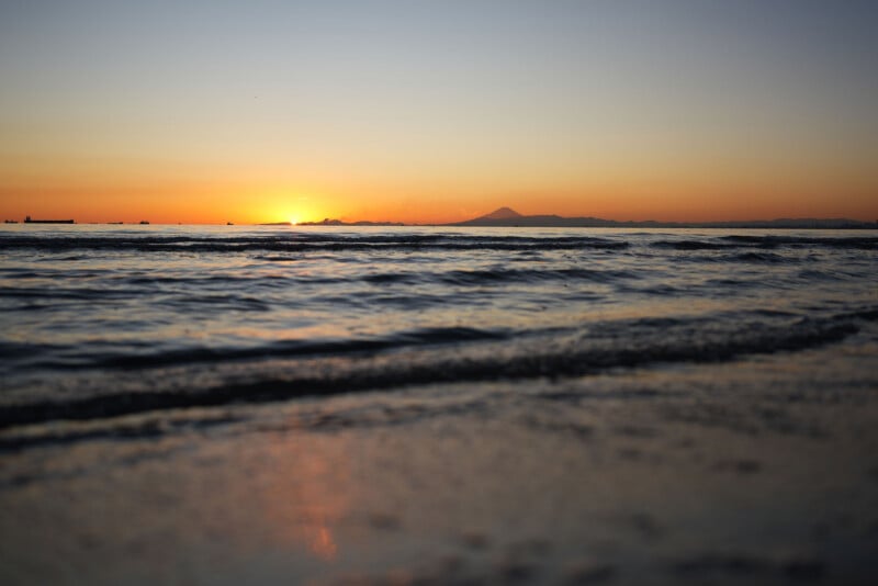 A serene beach scene at sunset, with gentle waves in the foreground and the sun setting behind distant mountains under a clear sky.