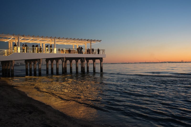 A group of people stand on a modern pier over calm water at sunset, with warm lights illuminating the structure and the sky glowing in shades of orange and blue. City lights are visible in the distant background.