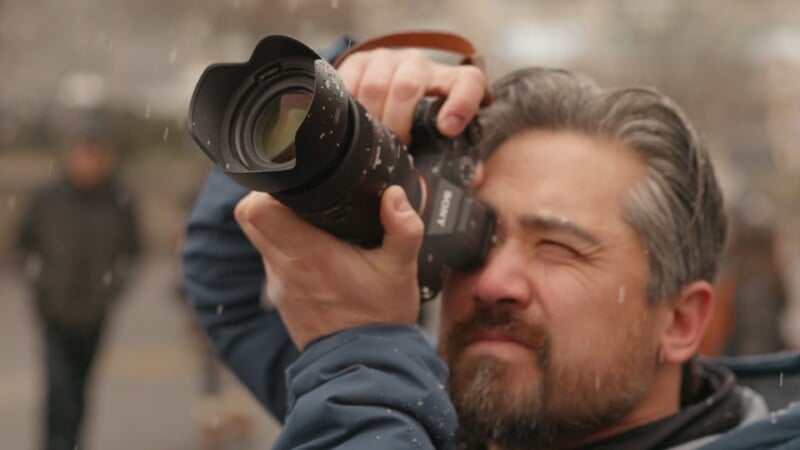A man with salt-and-pepper hair and beard is outdoors, holding a camera up to his face and squinting through the viewfinder as he takes a photo. Snowflakes are falling around him, and his expression is focused.