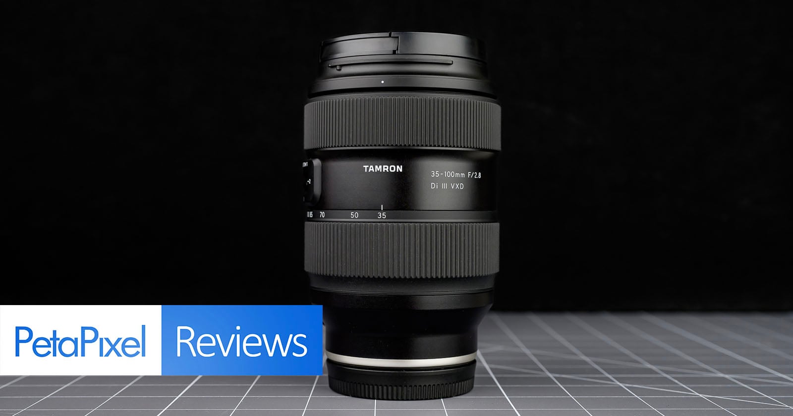 A Tamron 35-150mm F/2-2.8 camera lens stands upright on a grid-patterned surface with a black background. “PetaPixel Reviews” appears in blue and white at the bottom left corner.