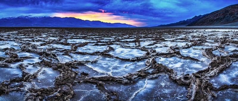 A dramatic sunset lights up the sky over a vast salt flat, featuring cracked, geometric salt formations. Mountains are visible in the background under a blue and orange sky.