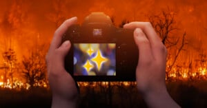 A person holds a camera, aiming at a forest fire. The background shows burning trees and orange flames, but the camera screen displays a blurred image of bright, glowing star shapes instead of the fire.