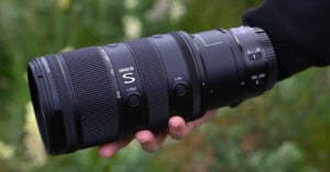 A hand holds a large NIKKOR S camera lens outdoors, with blurred green foliage in the background. The lens features visible buttons and detailed grip textures.
