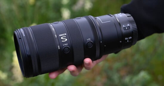 A hand holds a large NIKKOR S camera lens outdoors, with blurred green foliage in the background. The lens features visible buttons and detailed grip textures.