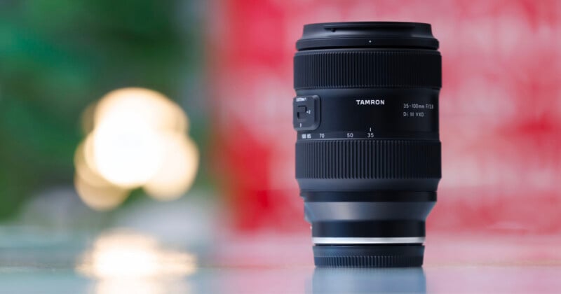 A Tamron 35-150mm camera lens is standing upright on a reflective surface, with a blurred green and red background and soft, circular light bokeh on the left side.