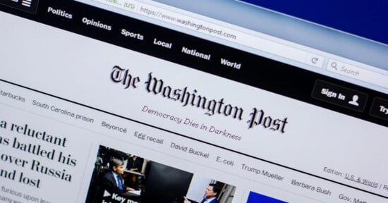 A computer screen displays the Washington Post website, showing the site’s logo, the slogan “Democracy Dies in Darkness,” and navigation links for sections like Politics, Opinions, Sports, and Local.