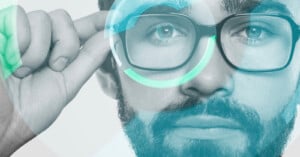 A close-up of a bearded person wearing glasses, touching the frame, with digital graphics overlaid around the glasses, suggesting technology or augmented reality.