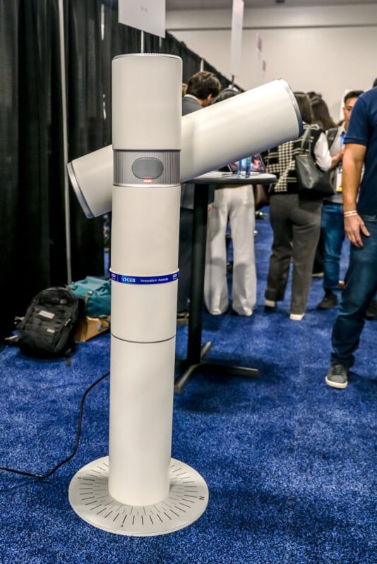A tall, modern, cylindrical air purifier with two angled side vents is displayed on a blue carpeted floor at an indoor event, surrounded by people and exhibition booths.