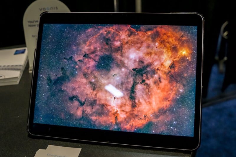 A tablet on a table displays a vibrant, high-resolution image of a reddish-orange nebula with black and white clouds in space. The background is dark and some paper items are partially visible nearby.