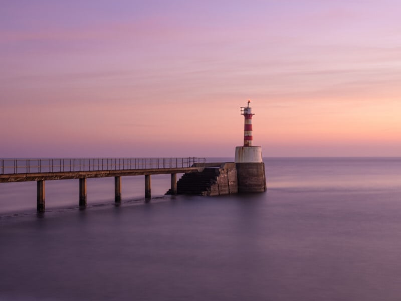 A red and white striped lighthouse stands at the end of a pier extending into calm water, with a pastel pink and purple sky at sunset or sunrise in the background.