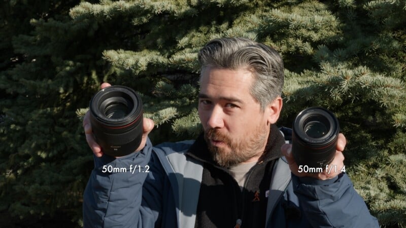 Un hombre parado frente a pinos al aire libre sosteniendo dos lentes de cámara etiquetados "50mm f/1.2" en su mano izquierda y "50mm f/1.4" en su mano derecha.