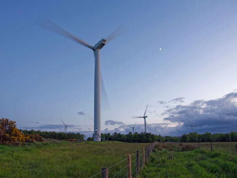 Una turbina eólica se encuentra en una pradera al anochecer, con otras turbinas al fondo y una luna creciente visible en el cielo despejado. Alambre de púas corre a lo largo del lado derecho de la imagen.