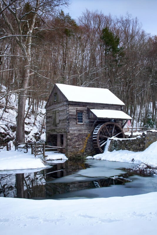 A rustic wooden watermill with a large waterwheel sits beside a partially frozen pond, surrounded by snow-covered ground and leafless trees in a winter landscape.