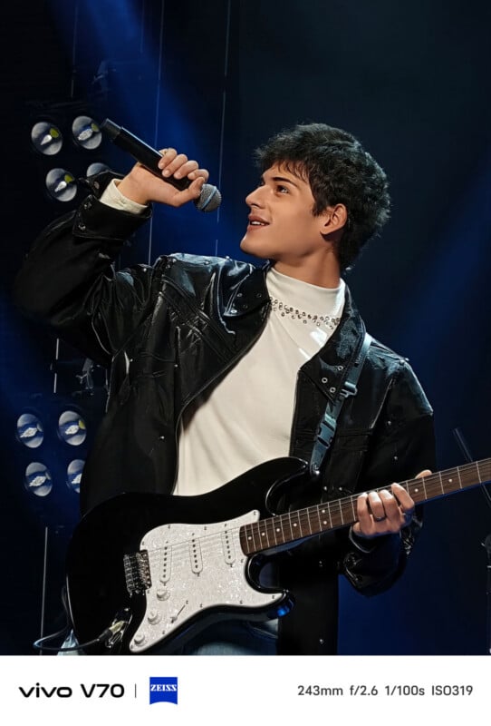 A young man wearing a black leather jacket and white shirt sings into a microphone and plays a black electric guitar on stage, with bright spotlights shining behind him.