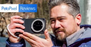 A man with gray hair and a beard holds a digital camera outdoors, aiming it to take a photo. The image has a "PetaPixel Reviews" logo in the top left corner. The background is blurred with natural, outdoor scenery.