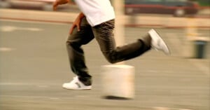 A person in a white shirt, dark jeans, and white sneakers is captured mid-run on a paved surface, with the background blurred to show motion.