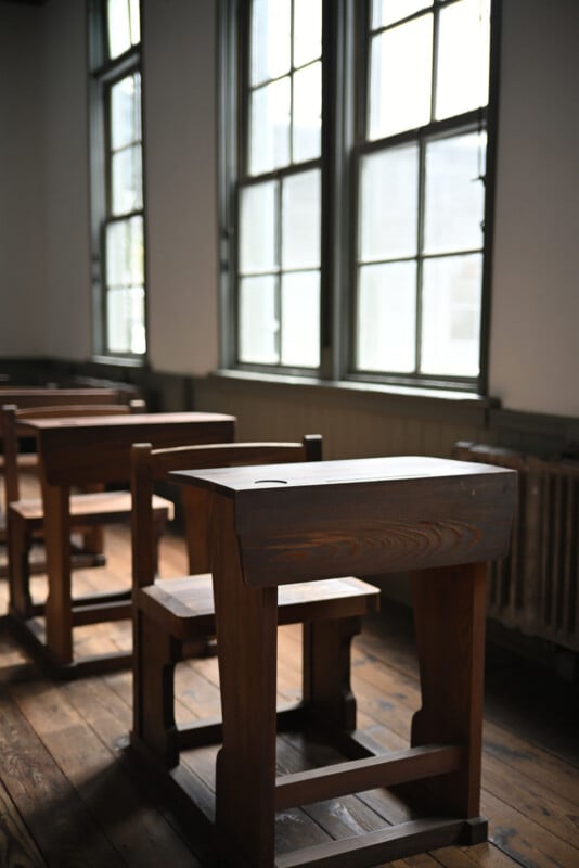 El aula tiene una hilera de mesas y bancos de madera vacíos, y grandes ventanales dejan entrar luz natural; La habitación tiene suelos de madera, lo que le da un aire retro y antiguo.
