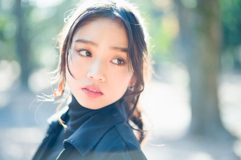 A young woman with long dark hair and soft makeup looks to the side outdoors. She wears a black jacket and turtleneck, with sunlight and blurred trees in the background creating a dreamy effect.