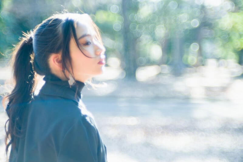 A young woman with long dark hair in a ponytail stands outdoors, looking over her shoulder. She wears a dark jacket. The background is brightly lit and blurred with circular bokeh effects.