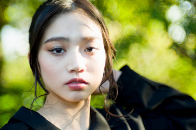 A young woman with long dark hair and natural makeup stands outdoors, wearing a black top. She looks at the camera with a calm expression, with sunlight and green foliage blurred in the background.