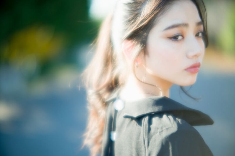 A young woman with long hair in a ponytail looks over her shoulder, wearing a black top. The photo has a soft, dreamy focus with bright natural light and a blurred outdoor background.