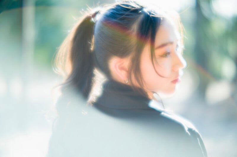 A young woman with long dark hair in a ponytail, wearing a black top, is looking over her shoulder in soft, dreamy sunlight with a rainbow lens flare in the background.