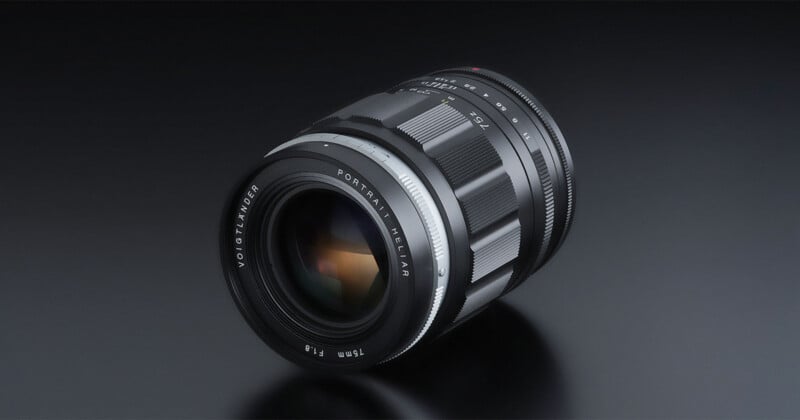 A Voigtländer camera lens with a metallic finish lies on a reflective dark surface, showing engraved text, ridged grip rings, and a prominent glass element.