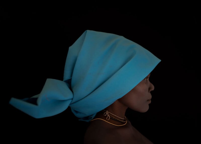 A person in profile against a black background wears a large, bright blue headwrap and gold necklaces. The lighting highlights their face and the texture of the fabric.