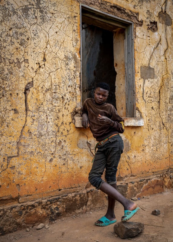 A boy leans against the windowsill of a weathered, cracked yellow wall, one hand resting on the sill, one foot crossed in front of the other. He wears a brown shirt, black pants, and blue-green sandals.
