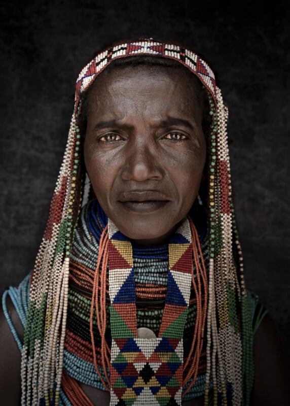 A woman wearing colorful, intricate beaded jewelry and headpiece stares directly at the camera, her expression calm and serious against a dark background.
