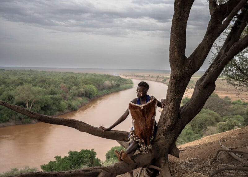 A person in traditional attire sits on a large tree overlooking a wide river and lush green landscape under a cloudy sky.