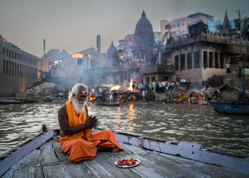 An elderly man in orange robes sits cross-legged on a wooden boat, hands in prayer, with a plate of offerings in front. Behind him, people gather along the riverbank with temples, fires, and smoke rising in the background.
