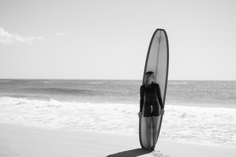 A person stands on a sandy beach, holding a tall surfboard upright beside them, with gentle waves and the ocean in the background. The image is in black and white.
