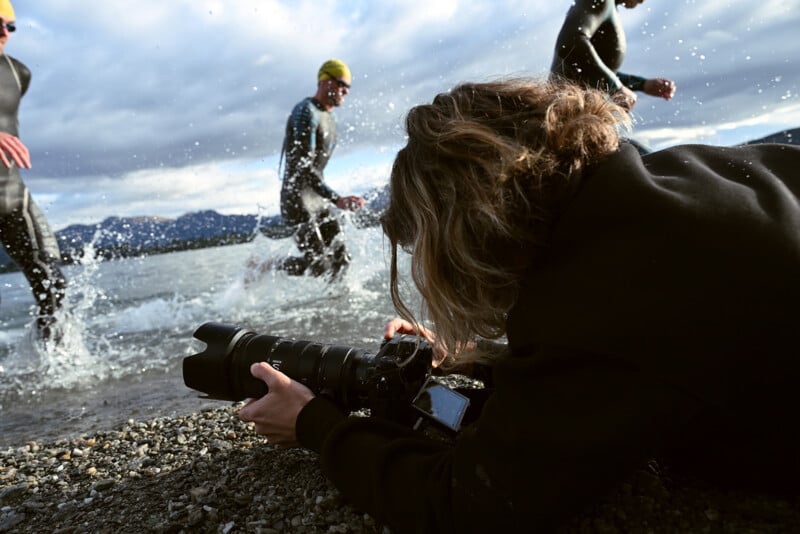 A photographer lying on pebbles by the water’s edge captures athletes in wetsuits and yellow caps running through shallow water during a race, with mountains and a cloudy sky in the background.