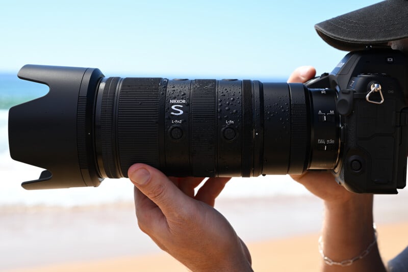 A person holding a Nikon camera with a large telephoto lens, showing water droplets on the lens, at a beach with the ocean and sand visible in the background.