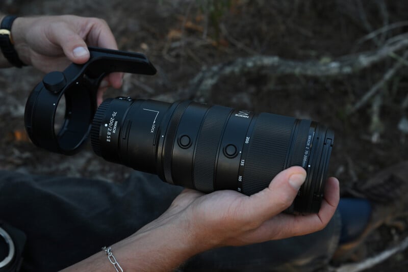 A person holds a large Nikon NIKKOR Z 180-600mm camera lens outdoors with one hand gripping the lens and the other holding a detachable tripod collar.