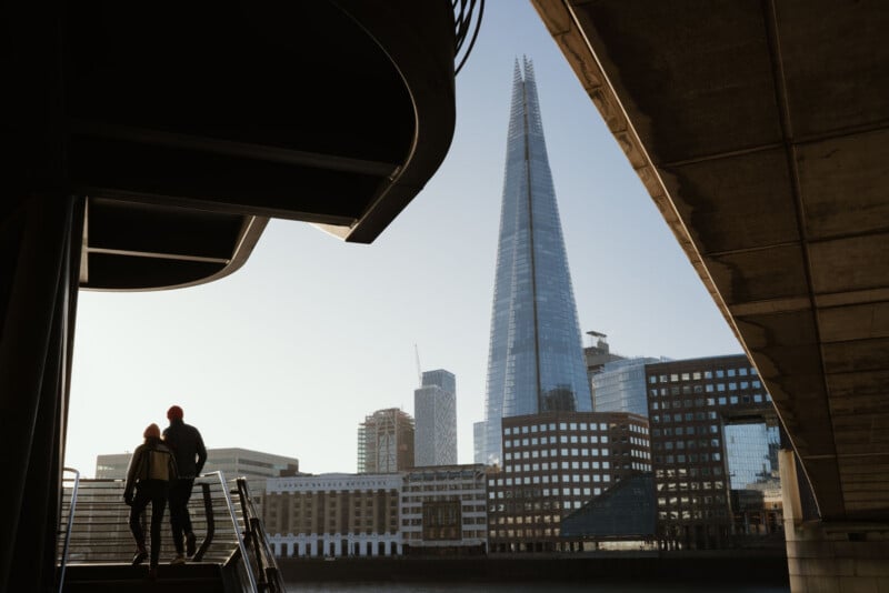 Two people stand at the top of outdoor stairs under a large structure, looking at the London skyline with The Shard skyscraper prominently in view on a clear day.