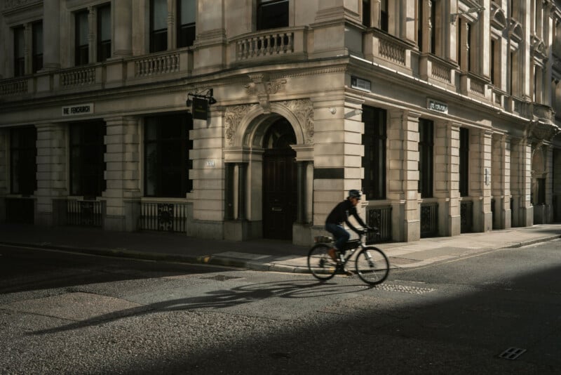 A person wearing a helmet rides a bicycle through a sunlit, empty city street, passing by an ornate, historic stone building with large windows and arched doorways.
