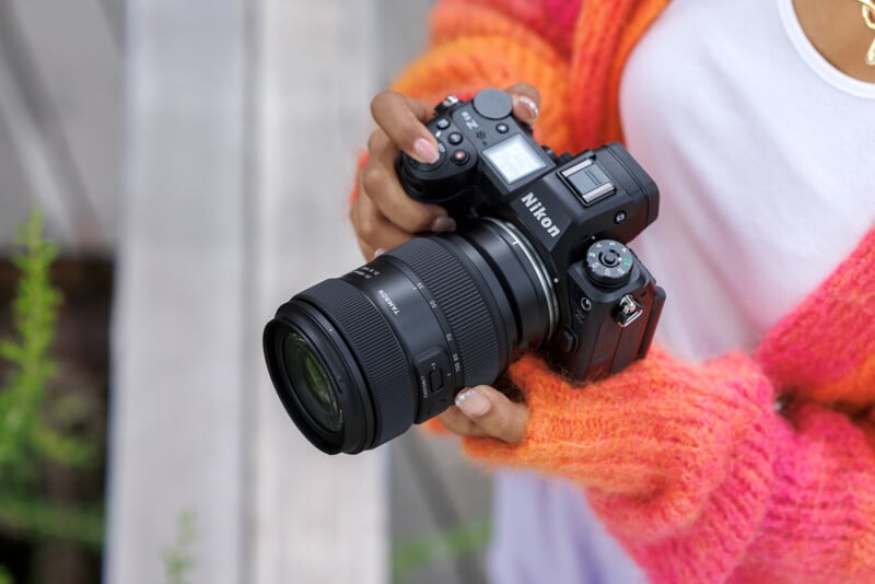 A person wearing a bright orange and pink sweater holds a Nikon mirrorless camera with a large lens, standing outdoors near a wooden surface.