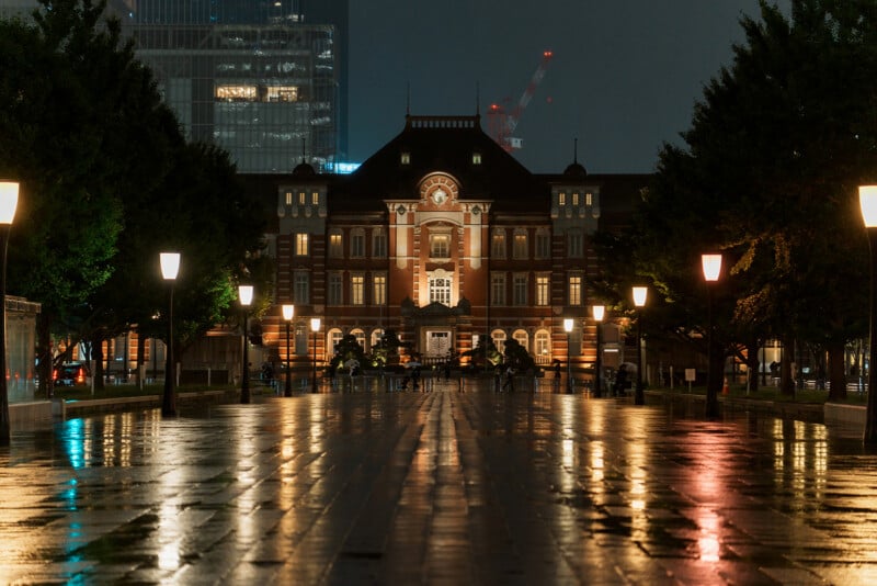 Tokyo Station at night, its historic red-brick facade illuminated. Wet pavement reflects streetlights and building lights, with trees lining both sides of the walkway leading to the station.