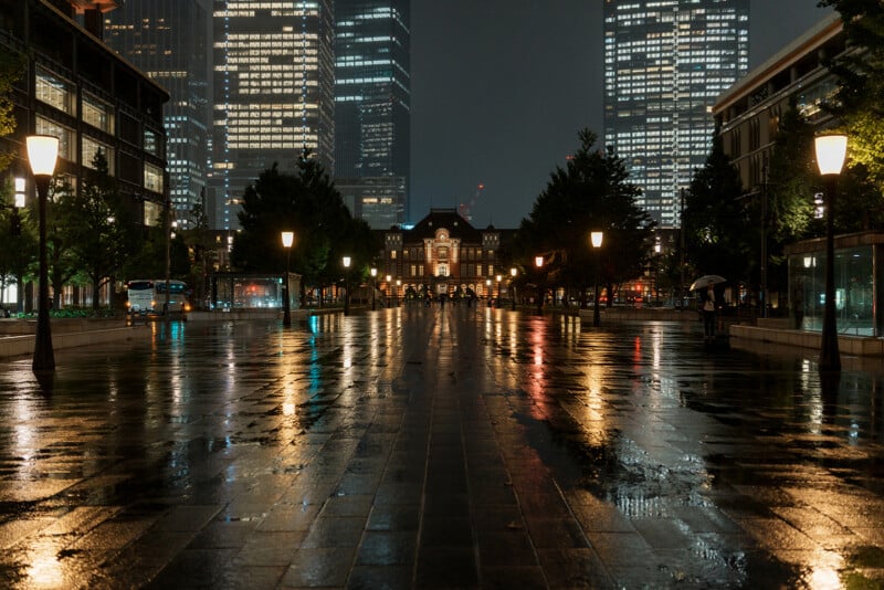 A wet, reflective street at night leads to a large, illuminated building. Skyscrapers and lit street lamps line both sides, with a few trees and people holding umbrellas visible in the scene.