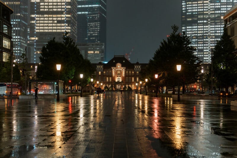 Tokyo Station at night, illuminated by streetlights and surrounded by tall modern skyscrapers, with wet pavement reflecting the city lights and a few people walking in the distance.